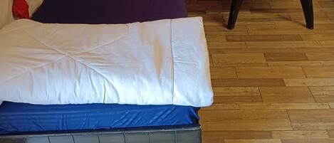 A bed with a white and blue mattress, a dark blue pillow, and red curtains. There is a small table with a box, a thermos, and glasses.