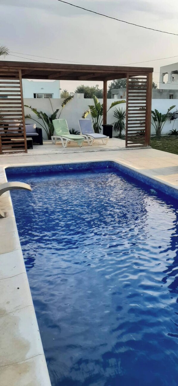 A rectangular swimming pool with a wooden pergola overhead, surrounded by lounge chairs and potted plants. - Luxurious Villa Rental in Houmt Souk, Djerba - Perfect Family Retreat (Houmt Souk)