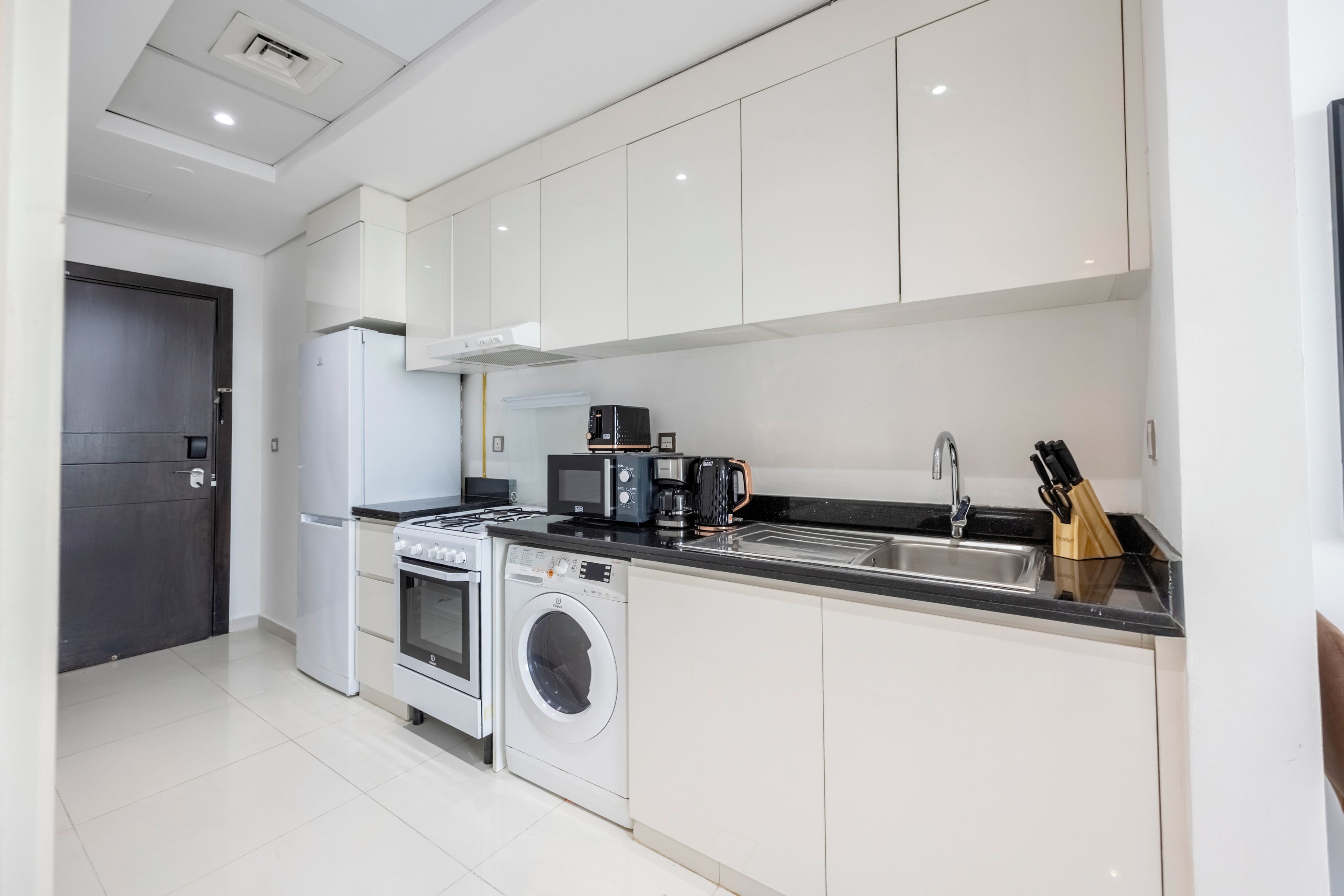 A modern kitchen with white cabinetry, a built-in oven, a washing machine, a microwave, and a coffee maker.