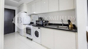 A modern kitchen with white cabinetry, a built-in oven, a washing machine, and a sink. - Bloomfields Cozy Stu Damac (Dubai)