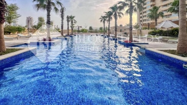 A swimming pool with clear blue water, surrounded by palm trees and a tall building in the background. - Bloomfields Cozy Stu Damac (Dubai)