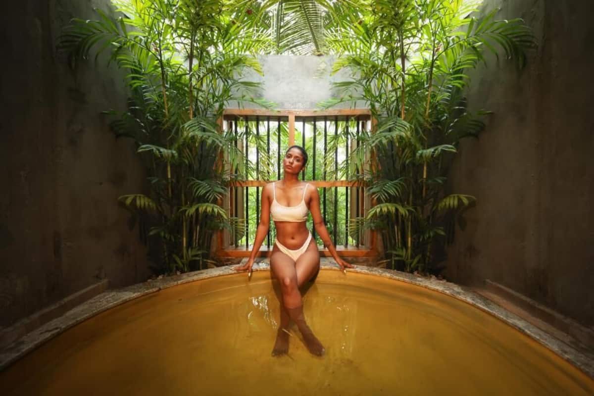 A person standing in a pool with a wooden structure and lush greenery in the background.