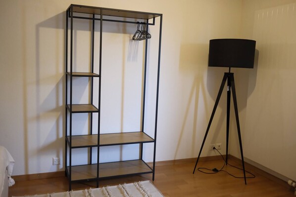 Une pièce avec une étagère en métal noir adossée à un mur, un lampadaire avec un abat-jour noir et un tapis blanc sur le plancher en bois.