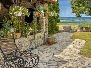 Un chemin de pierres mène à un bâtiment rustique, agrémenté de jardinières suspendues et d’un banc.