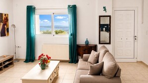 Ein Wohnzimmer mit Sofa, Couchtisch mit Blumen, Heizkörper, Fenster mit Vorhängen, Spiegel und Tür.