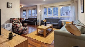 A modern living room with a wooden coffee table, floral armchair, grey sofa, and a view of the city through large windows. - Paris View & Direct Access  Apartment 10 people (Courbevoie)