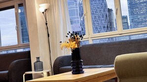 A modern living room with a wooden coffee table, a dark vase with flowers, a grey sofa, and a floor lamp. Large windows offer a view of city buildings. - Paris View & Direct Access  Apartment 10 people (Courbevoie)