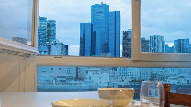 A dining table with a bowl and utensils, a glass, and a window with a cityscape view. - Paris View & Direct Access  Apartment 10 people (Courbevoie)