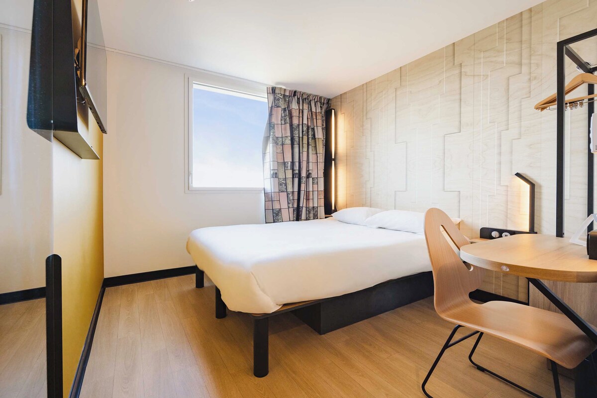 Habitación de hotel con cama, escritorio, silla, televisor y ventana con cortinas.