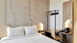 A hotel room with a bed, a desk with a chair, a hanging coat rack, and a bedside table with a lamp.
