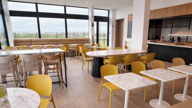 A modern dining area with yellow chairs and white tables, large windows with a view, and a kitchen area.