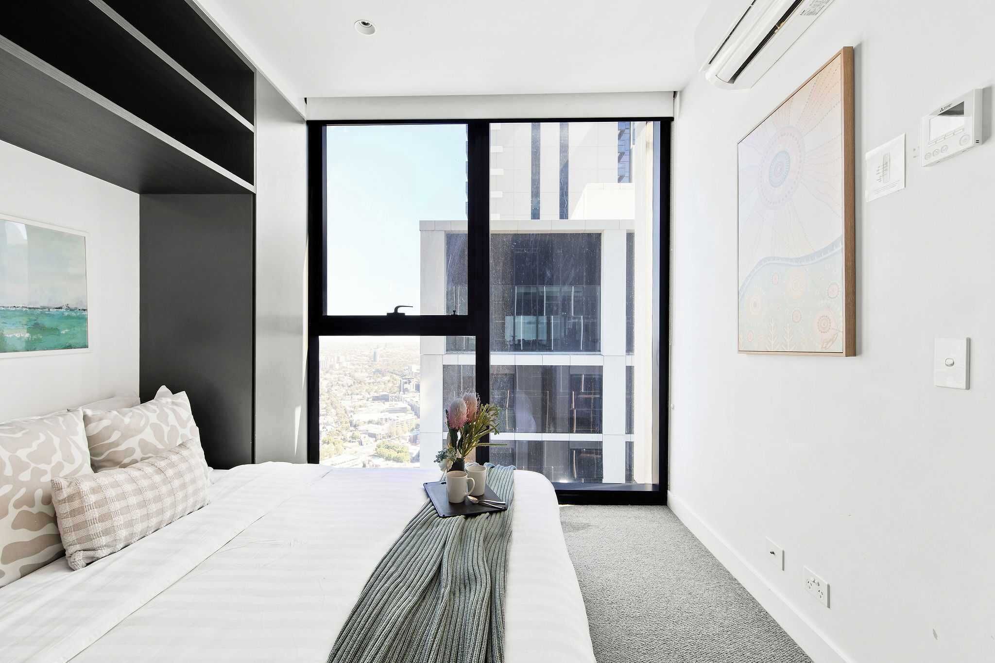 Een moderne hotelkamer met een groot raam, een bed met gestreepte kussens, een nachtkastje met een vaas en een mok, en een kunstwerk in een lijst aan de muur.