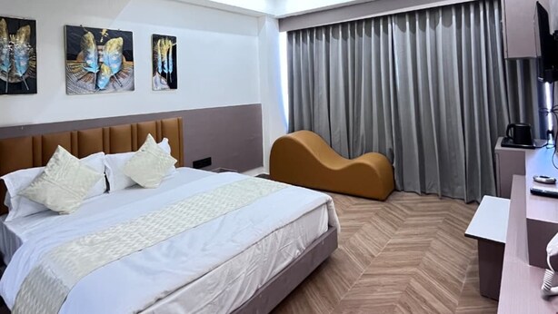 A modern hotel room with a large bed, a curved sofa, a desk with a lamp, and artwork on the walls. - Hotel Lee Prime (Ahmedabad)