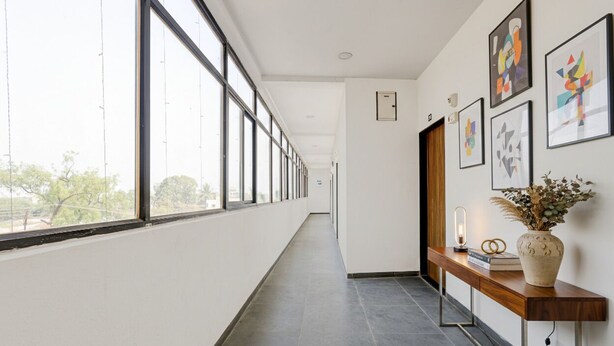 Un long couloir étroit, avec de grandes fenêtres d’un côté, un banc en bois orné d’un vase et d’un livre, et des œuvres d’art encadrées accrochées au mur.