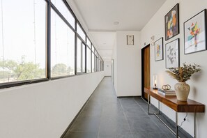 Un pasillo largo y estrecho con ventanas amplias en un lado, un banco de madera con un jarrón y un libro, y obras de arte enmarcadas en la pared.