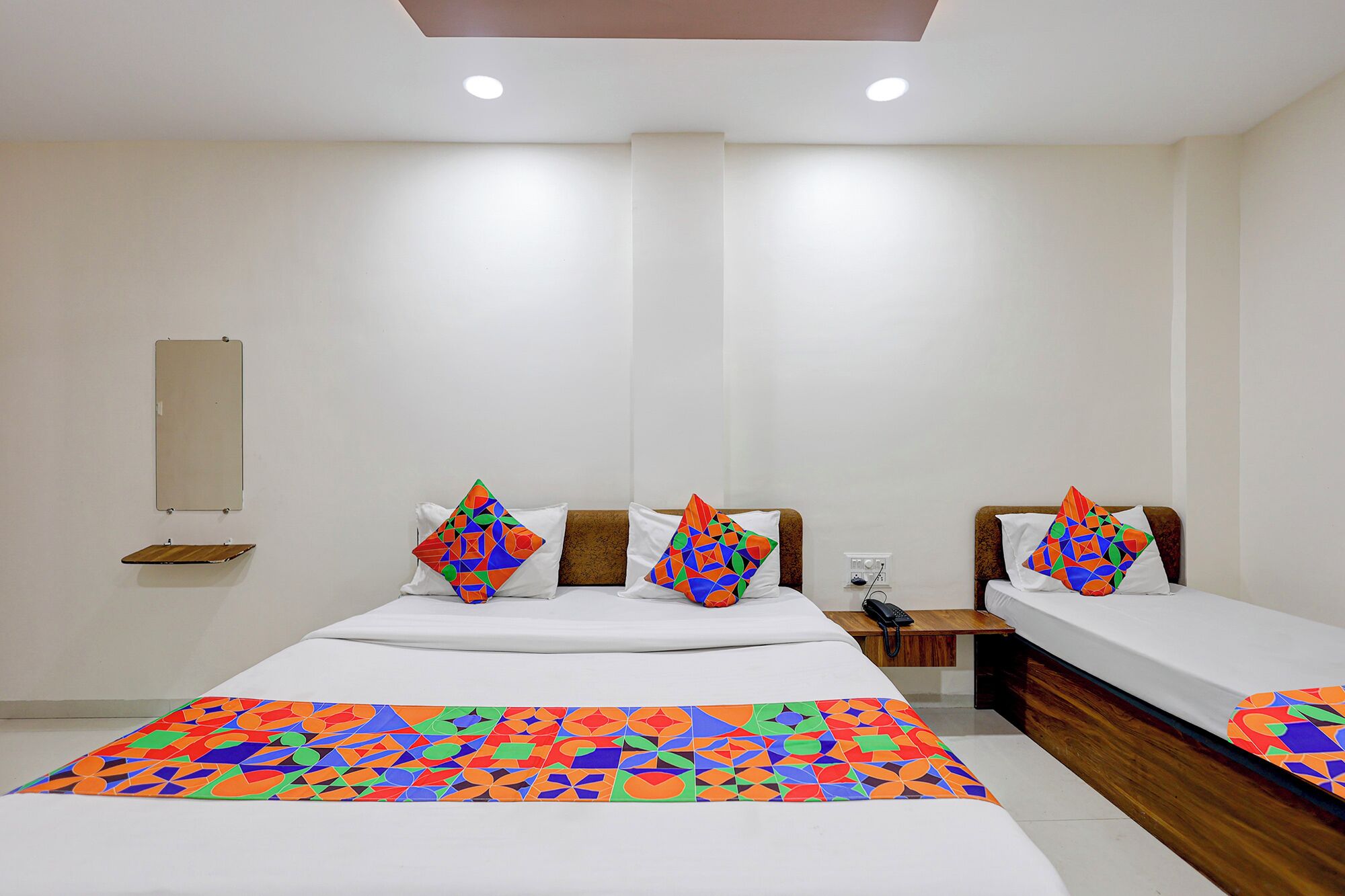Una habitación de hotel con dos camas, un cabecero de madera, una mesita de noche con un teléfono y una colcha de diseño geométrico y colores vivos.