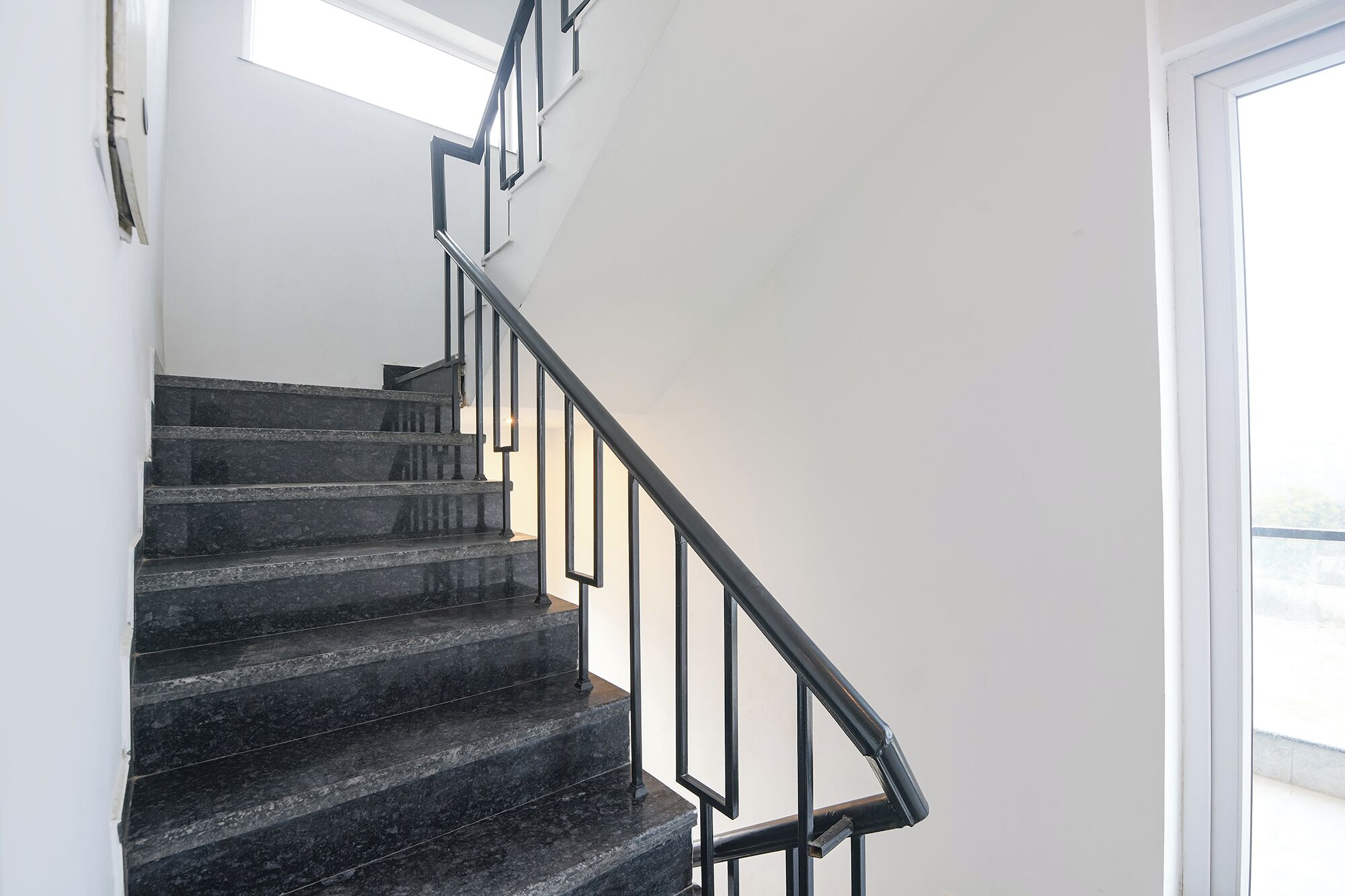 A staircase with black steps and metal railings, leading to a bright room with a white wall and a window.