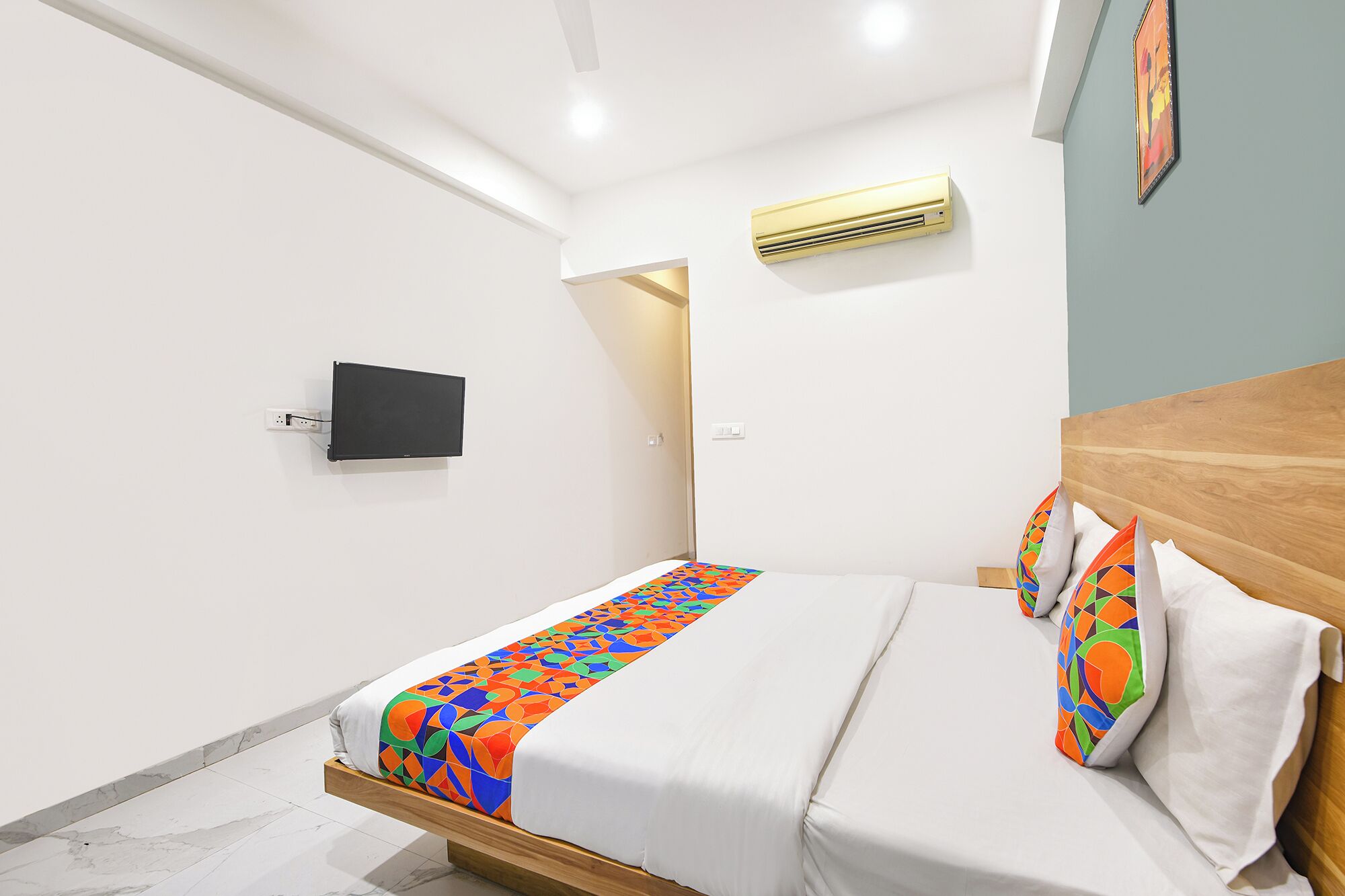A modern hotel room with a bed, a flat-screen TV, an air conditioning unit, and a door leading to another room.