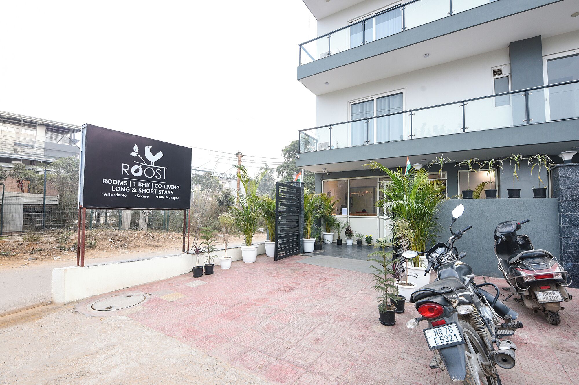 Un edificio moderno con un cartello pubblicitario per 'ROOST', che offre camere, co-living e soggiorni brevi. Sono presenti piante in vaso e due motociclette parcheggiate davanti.