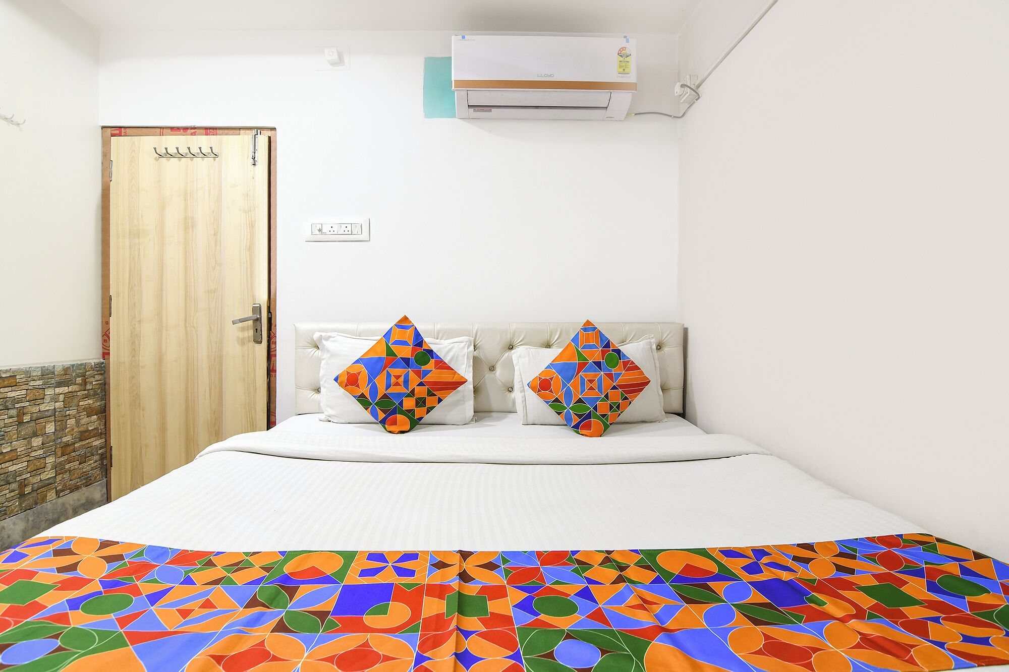 A bedroom with a bed, two pillows with geometric patterns, a wooden door, and an air conditioning unit on the wall.