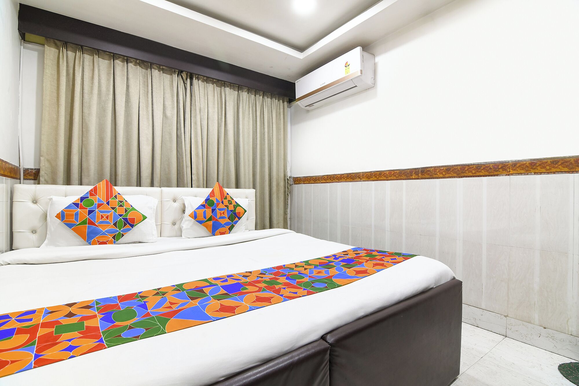 Una camera d'albergo con un letto, una finestra con tende, un condizionatore d'aria e un cuscino decorativo.