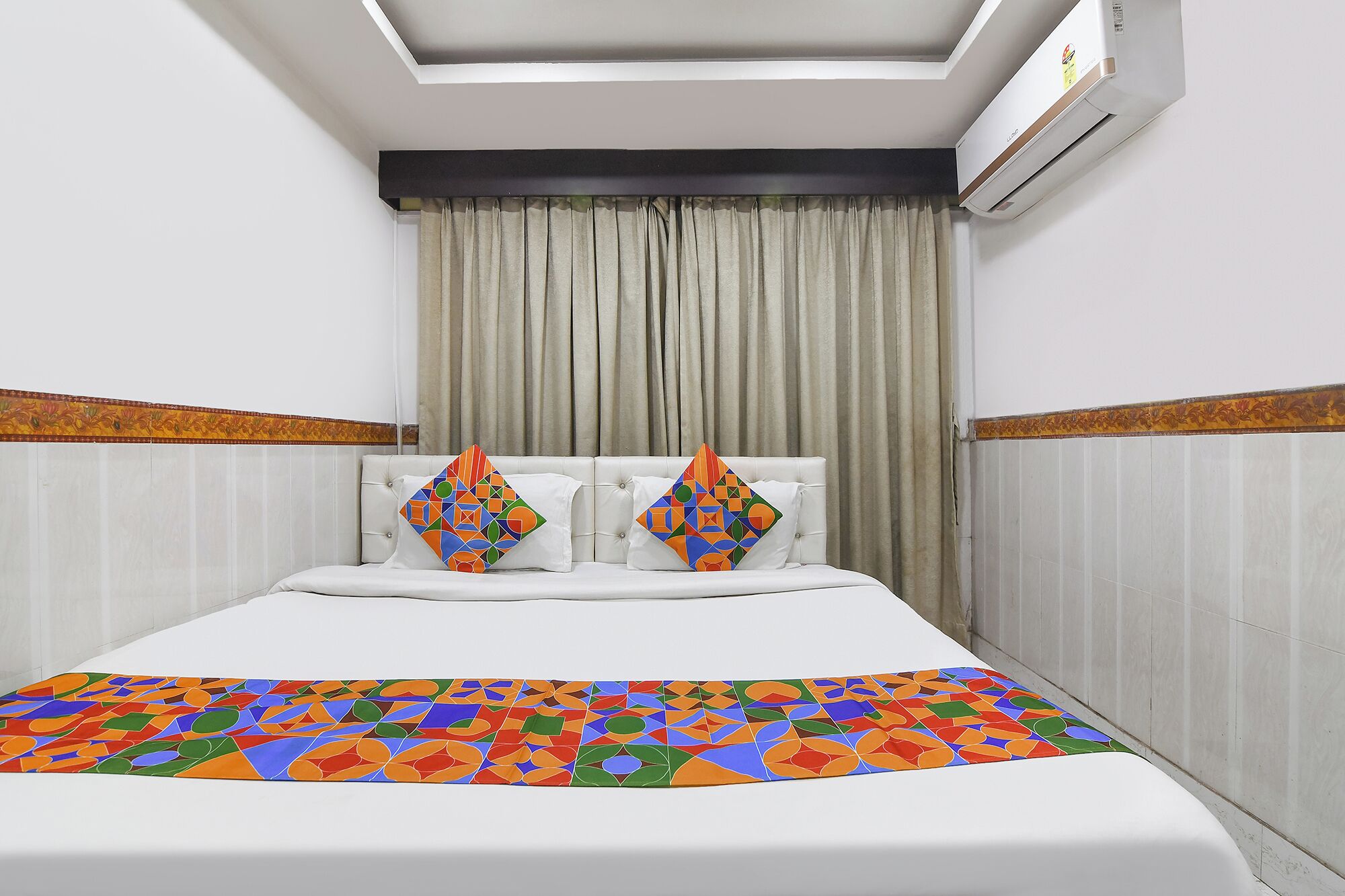 Une chambre d’hôtel avec un lit, deux oreillers aux motifs géométriques colorés, un ventilateur de plafond et une fenêtre avec des rideaux.