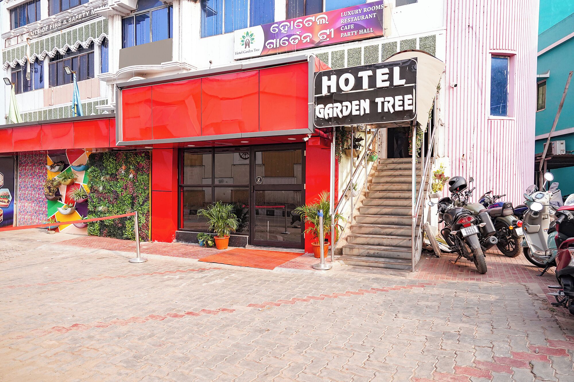 A hotel named "Hotel Garden Tree" with a red facade, a staircase leading to the entrance, and several motorcycles parked outside.