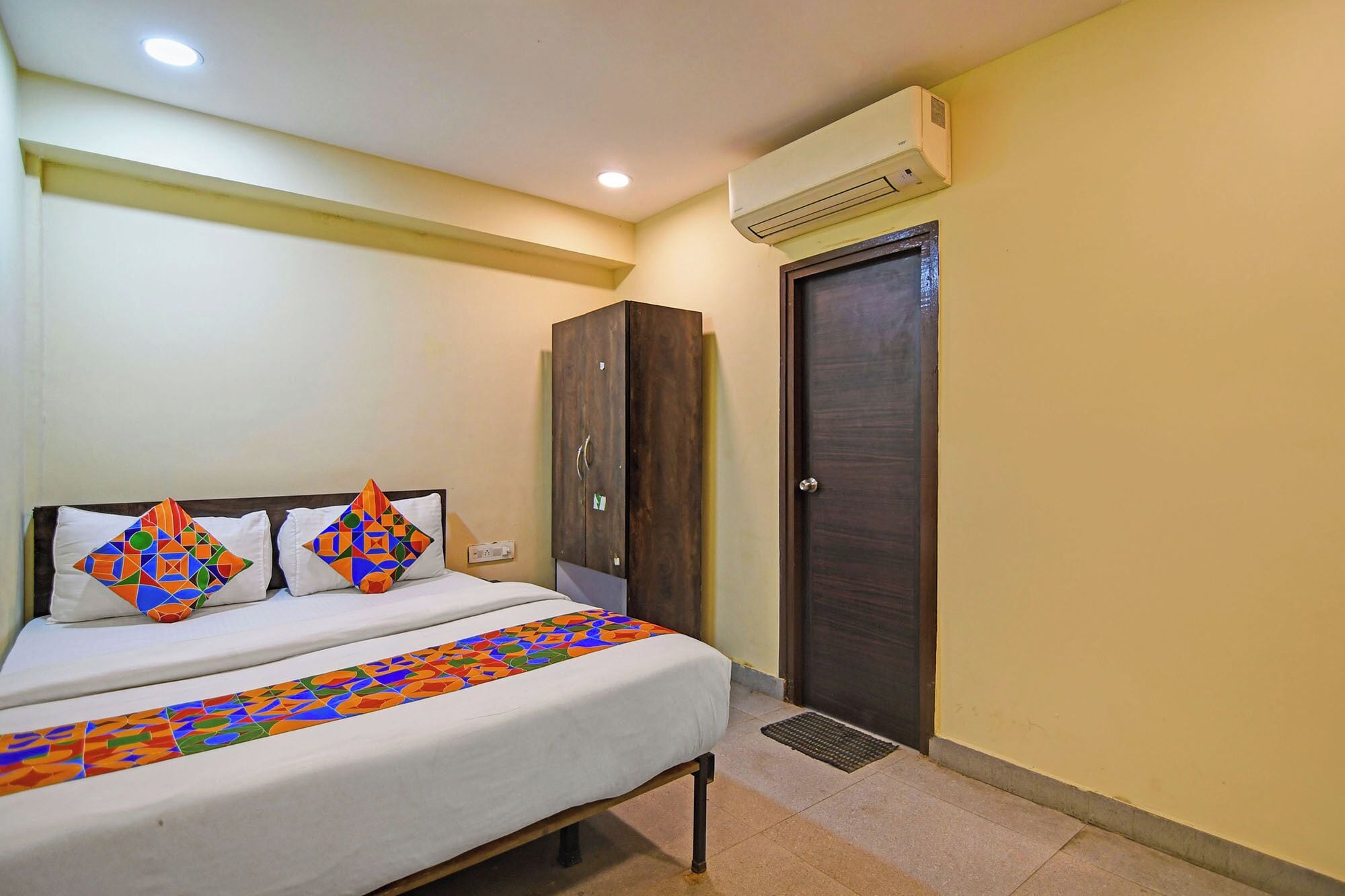 A hotel room with a bed, two pillows with colorful geometric patterns, a wardrobe, and an air conditioning unit.