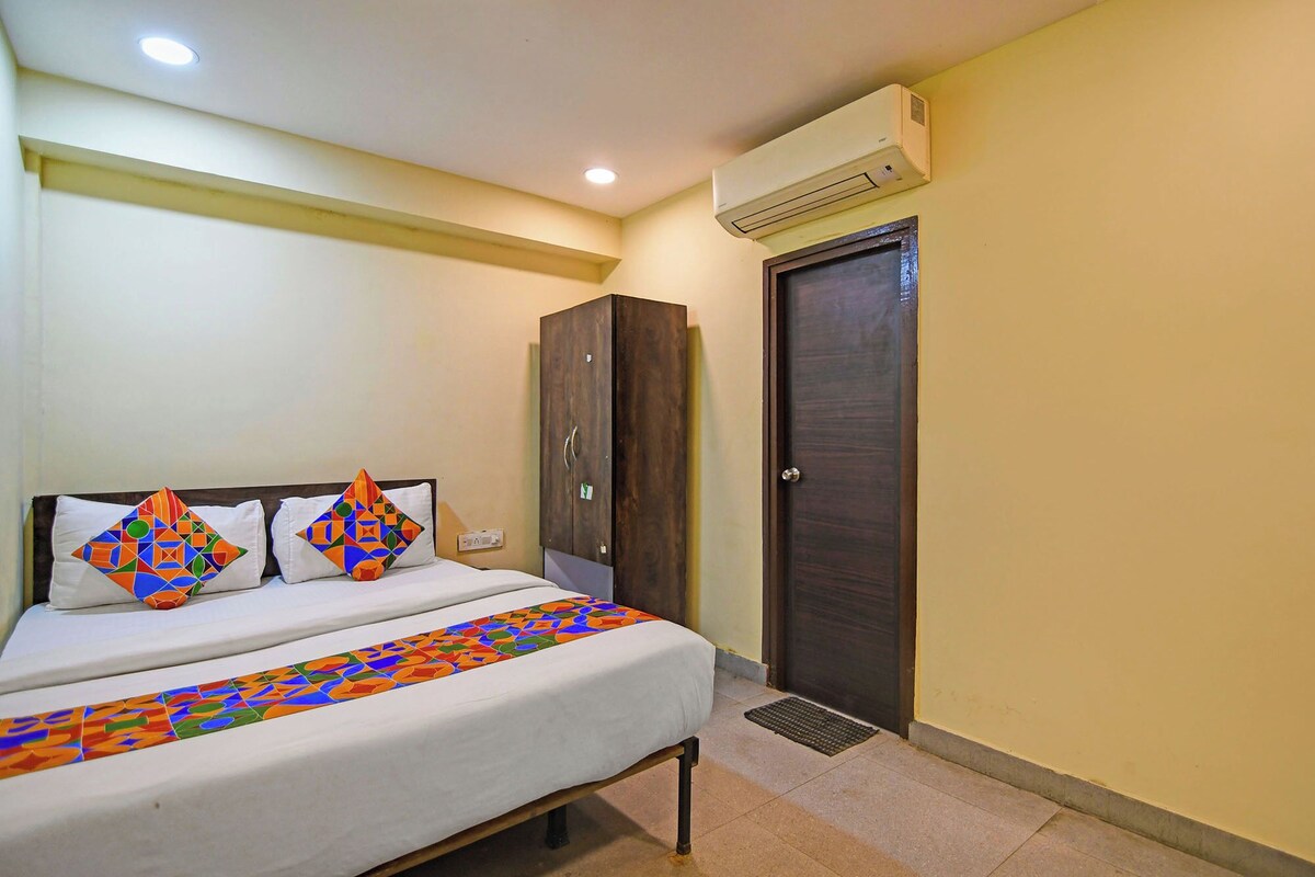 A hotel room with a bed, two pillows with colorful geometric patterns, a wardrobe, and an air conditioning unit.