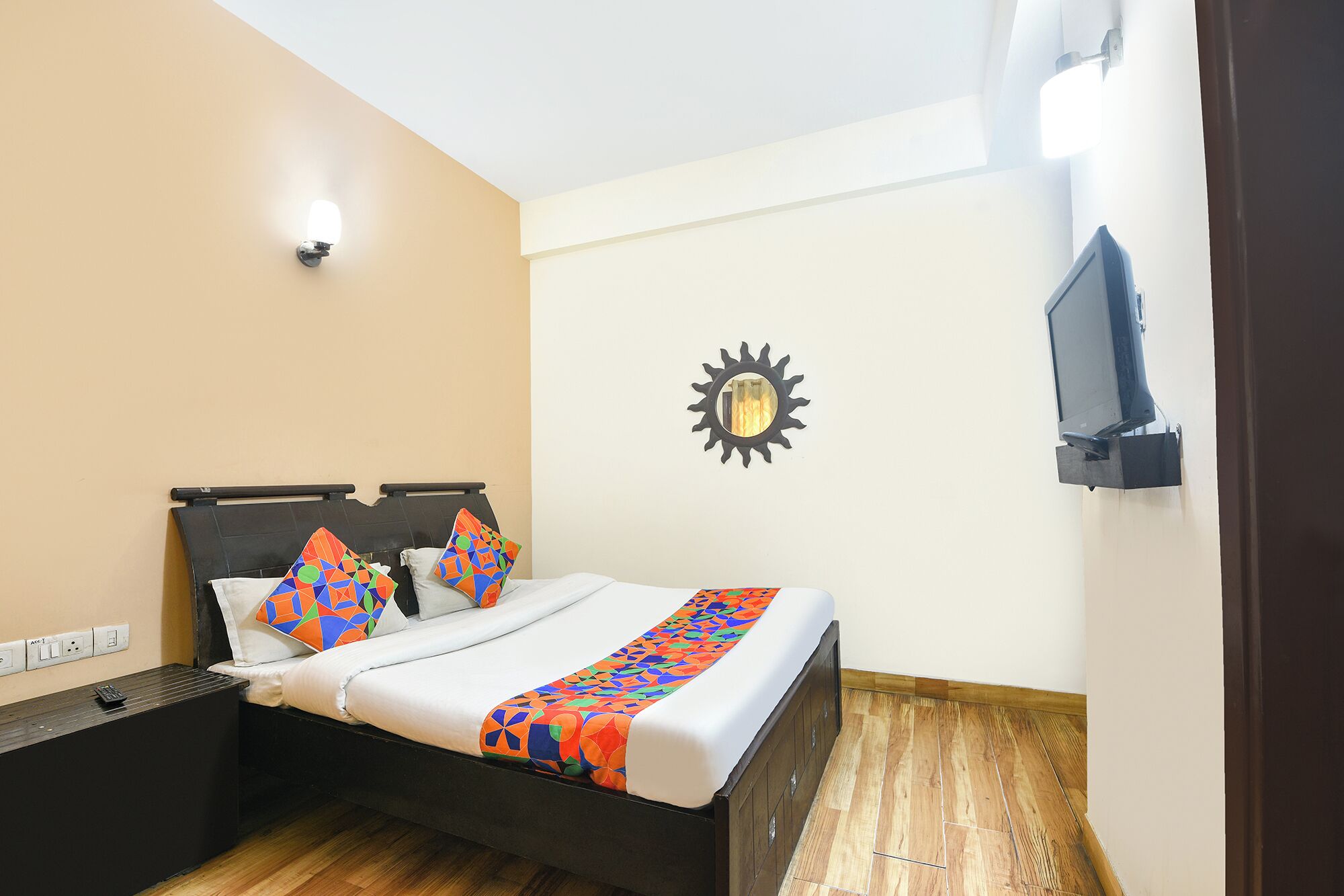 A neatly made bed with colorful pillows and a patterned bedspread, a wall-mounted TV, and a sun-shaped mirror.