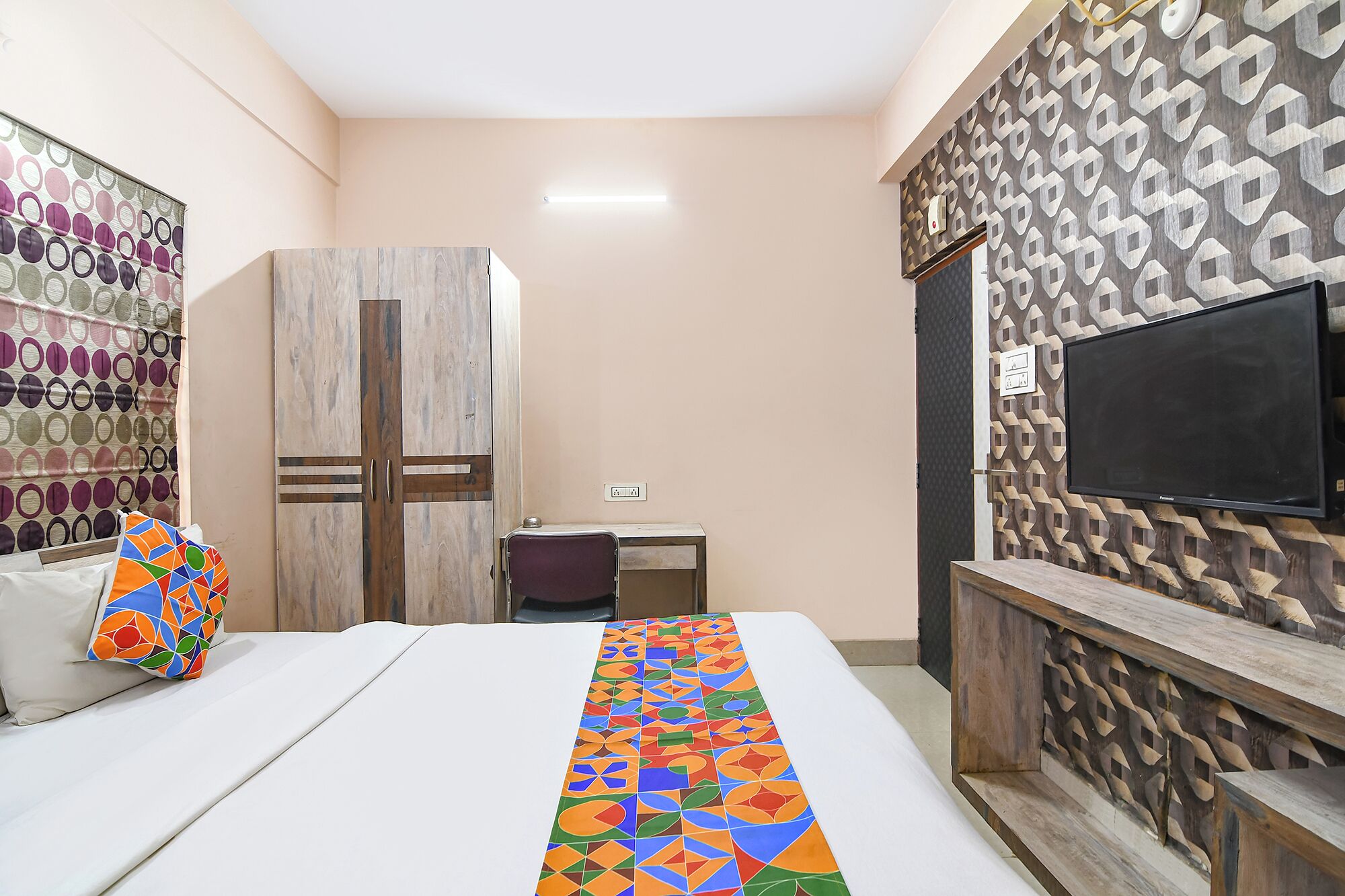 Ein Hotelzimmer mit einem Bett, einem Schreibtisch, einem Kleiderschrank und einem Fernseher an einer Wand mit geometrischem Muster.