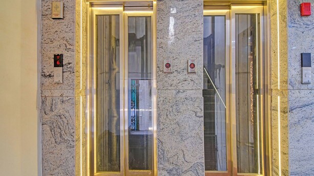 An elevator with marble walls and glass doors, emergency alarm buttons, and a red emergency phone on the wall. - Fabhotel Disa (Kolkata)
