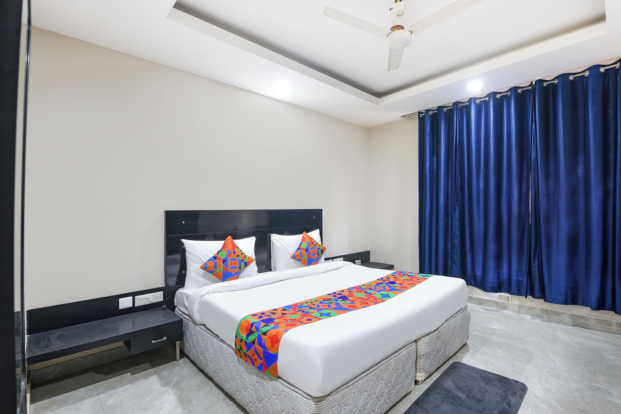 Une chambre d’hôtel bien rangée, avec un lit, une table de chevet, un ventilateur de plafond moderne et une fenêtre ornée de rideaux bleus.