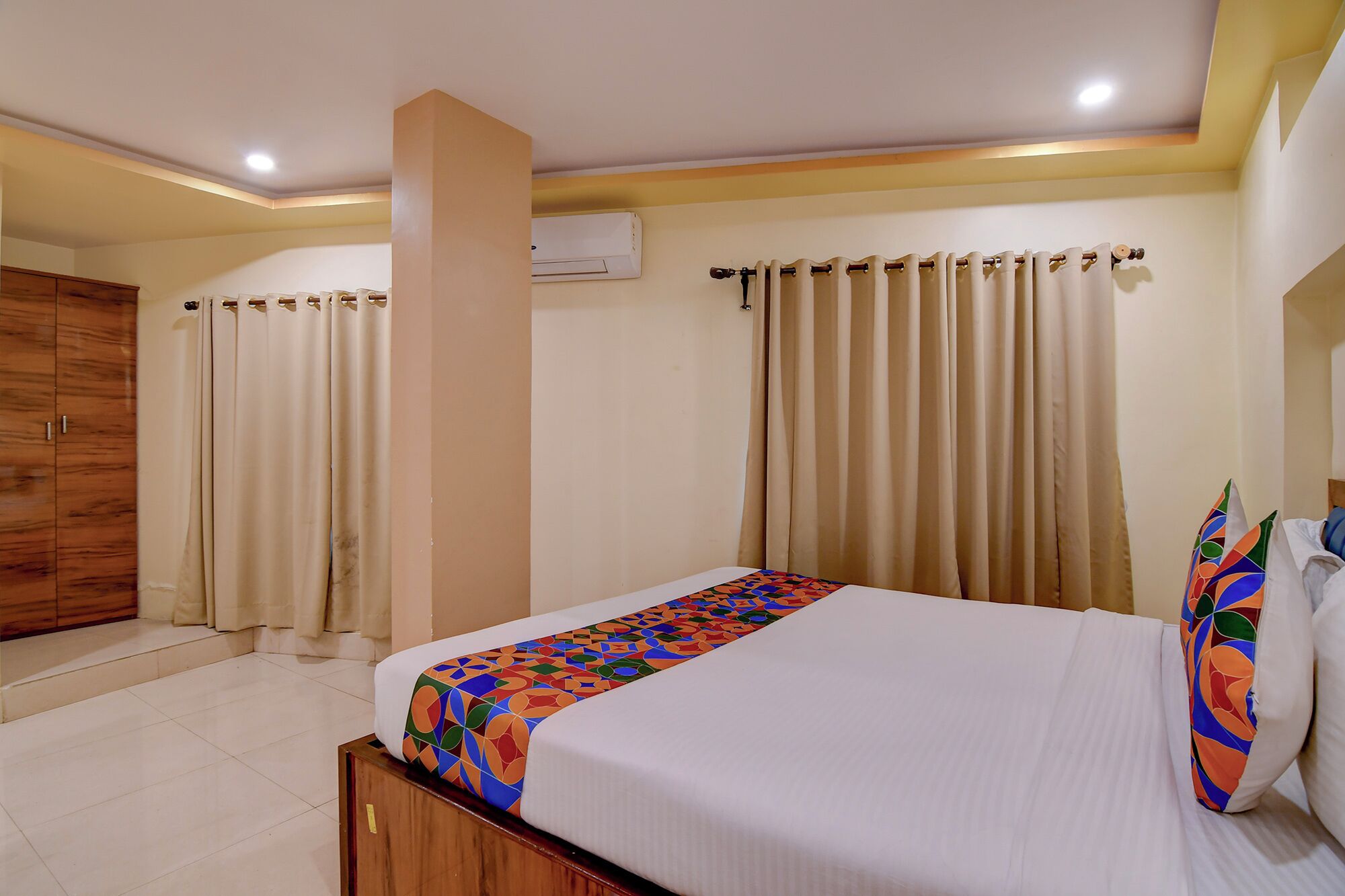 A hotel room with a bed, curtains, a wooden wardrobe, and a ceiling light.