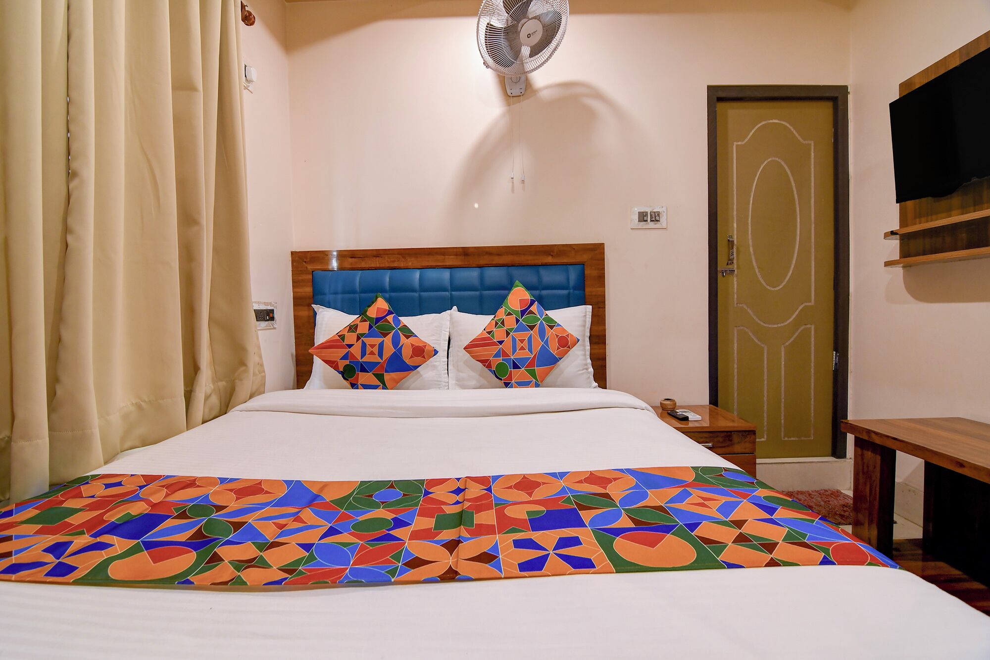 A hotel room with a bed, a nightstand, a fan, a door, and a TV on a shelf.