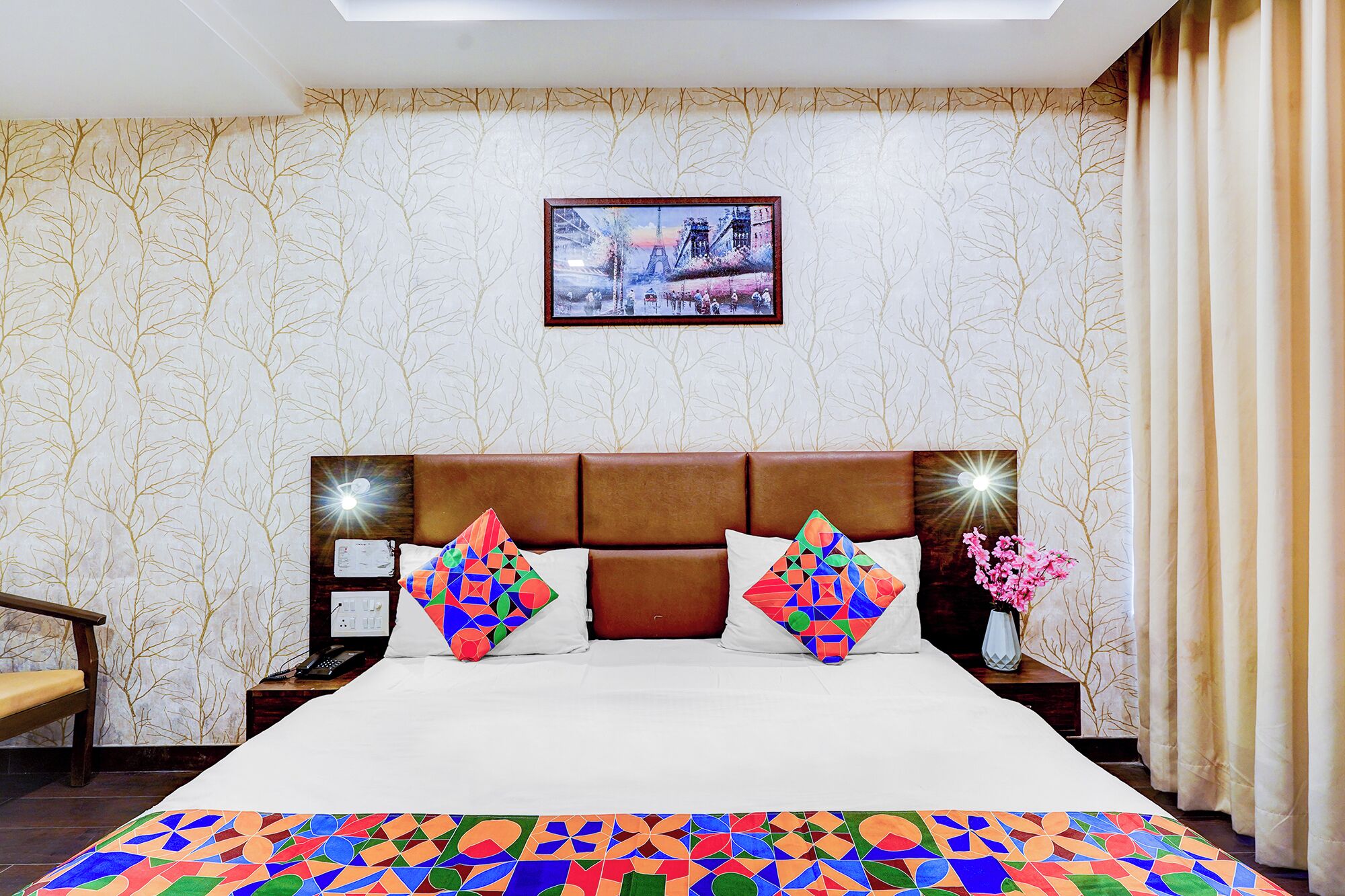 A hotel room with a large bed, a headboard with lights, a nightstand, a chair, and a wall with a framed picture.