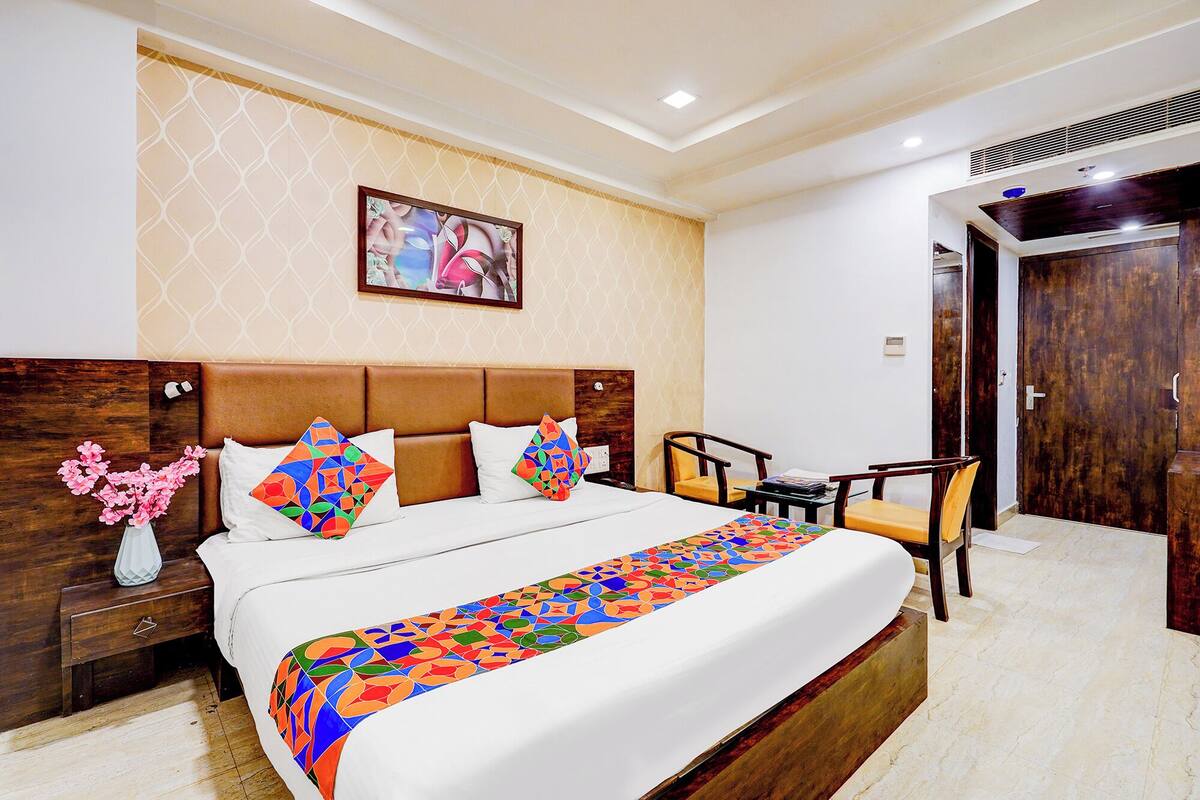 Habitación de hotel con una cama grande, cabecera de madera, una mesita de noche con un florero, una silla y una puerta.