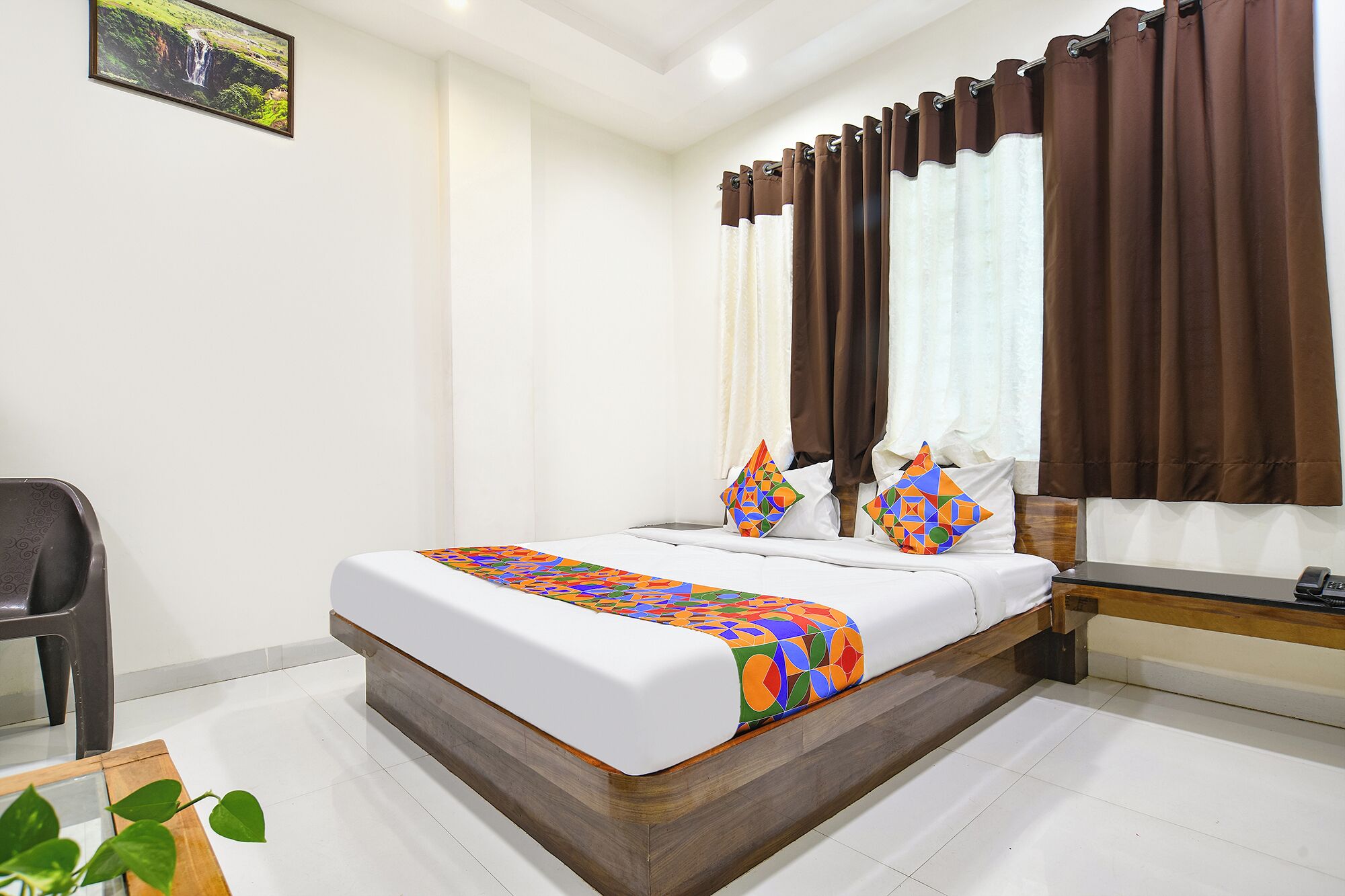 A neatly made bed with a colorful geometric-patterned bedspread, two pillows with a similar design, a wooden headboard, and a window with brown curtains.