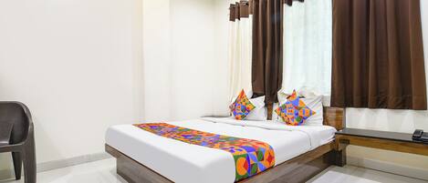 A neatly made bed with a colorful geometric-patterned bedspread, two pillows with a similar design, a wooden headboard, and a window with brown curtains.