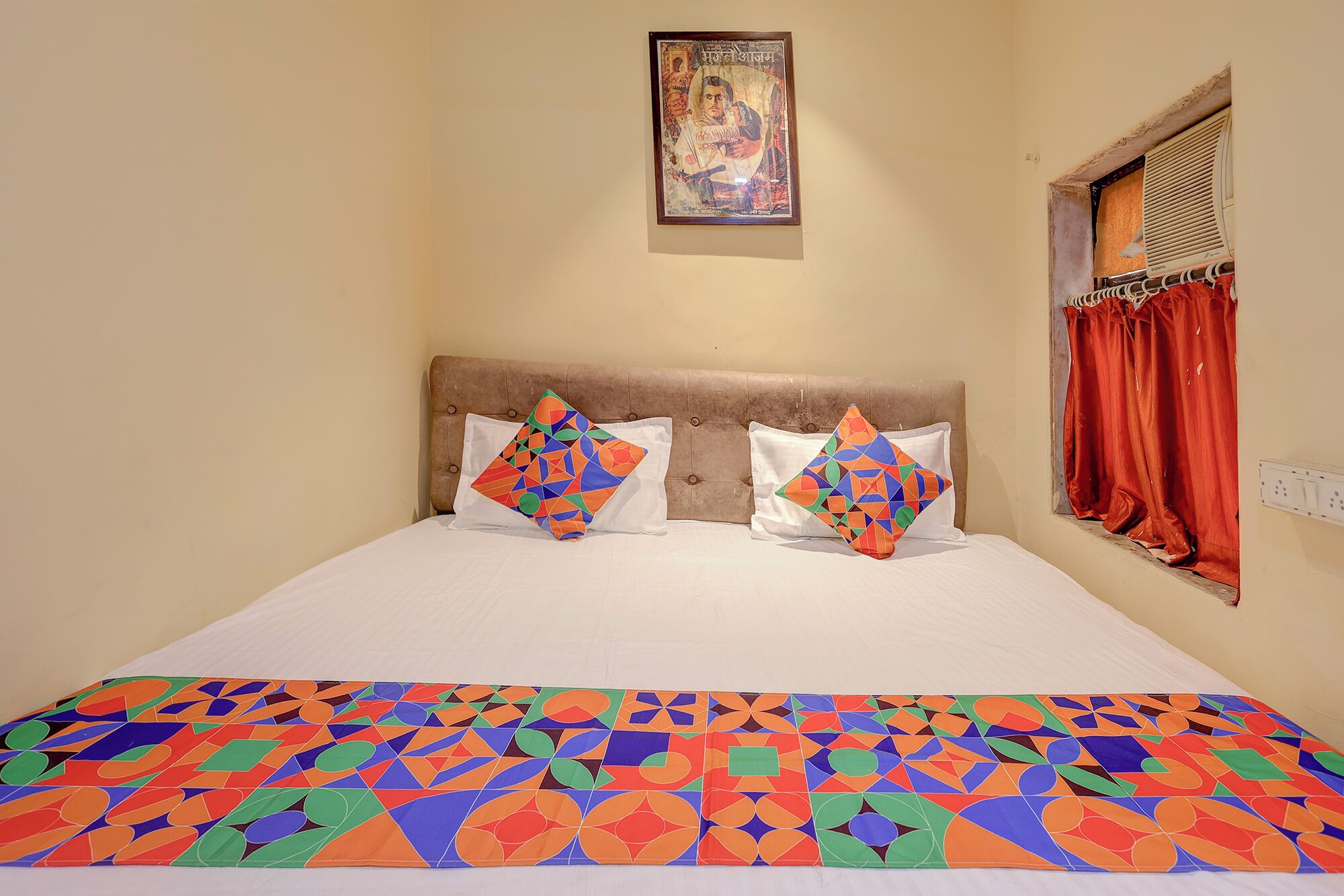 A bed with a patterned bedspread, two pillows with geometric designs, a headboard, a wall-mounted picture, and a window with red curtains.
