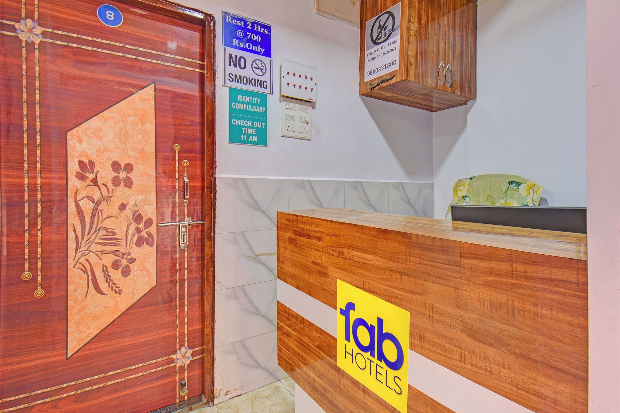 A hotel reception area with a wooden reception desk, a floral door, and various signs including a no smoking sign and a check-out time sign.