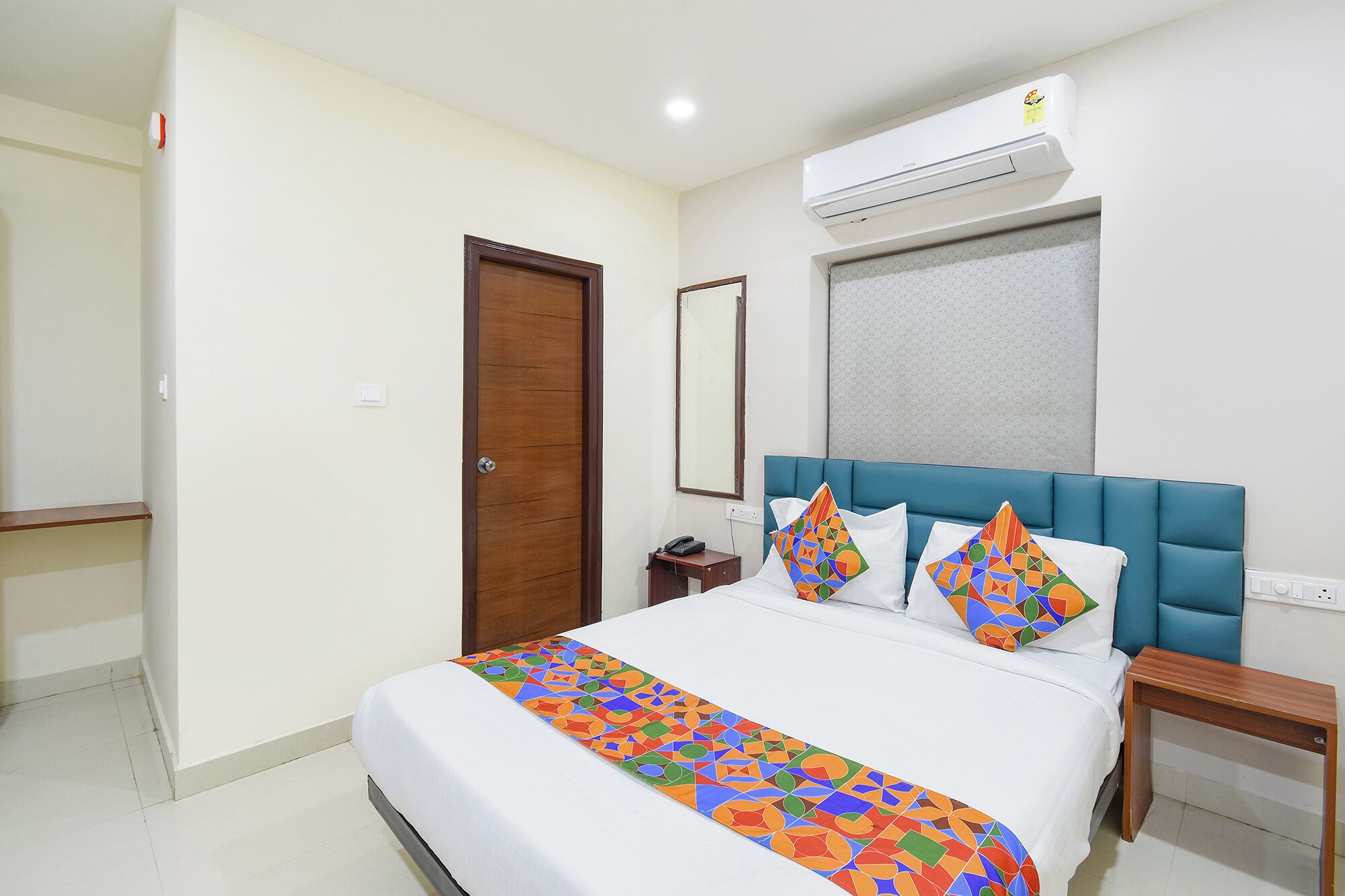 A hotel room with a bed, a nightstand, a wooden door, an air conditioning unit, and a window with blinds.