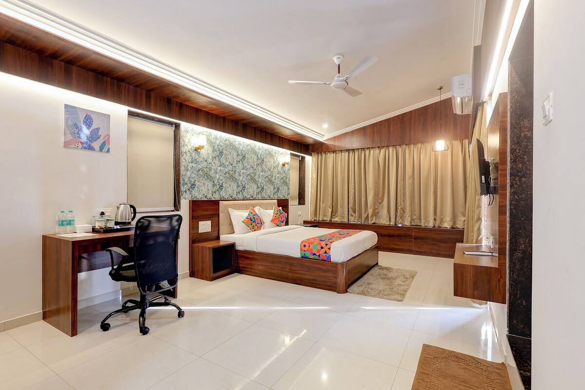 A modern hotel room with a wooden headboard, a desk with a chair, a bed with a colorful bedspread, a ceiling fan, and a large window with curtains.
