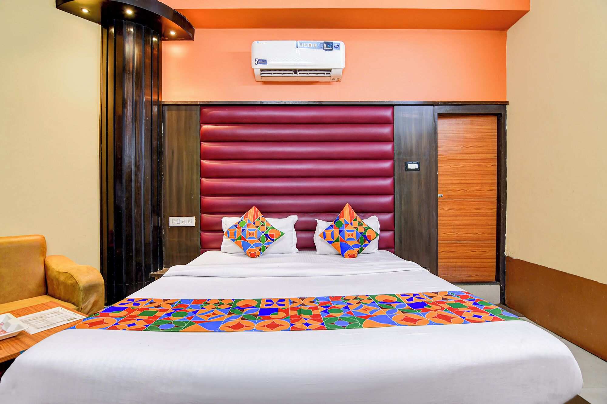 A hotel room with a bed, a red upholstered headboard, a wooden door, and an air conditioning unit on the wall.