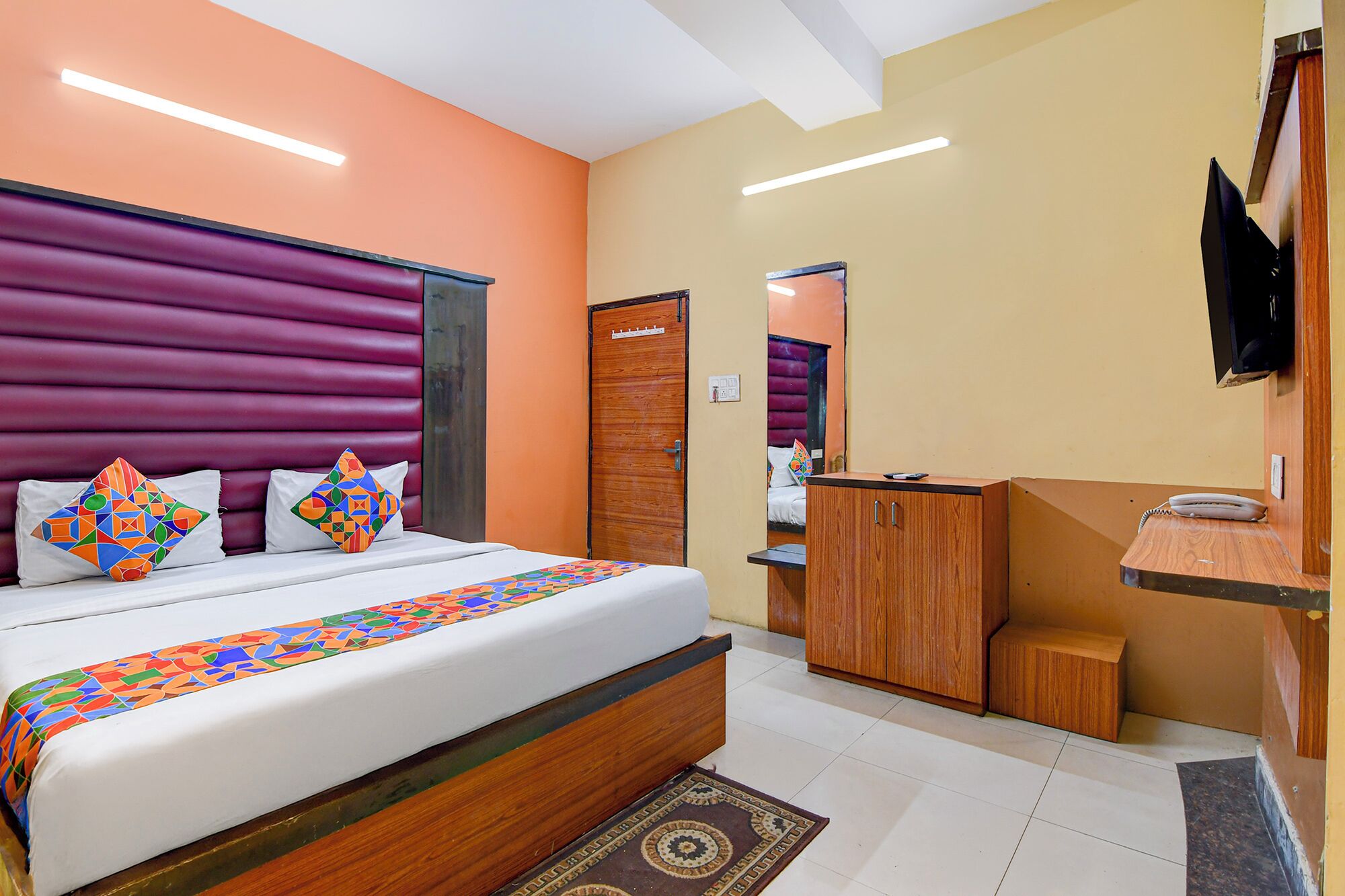 A hotel room with a bed, a wooden wardrobe, a desk with a telephone, and a television mounted on the wall.
