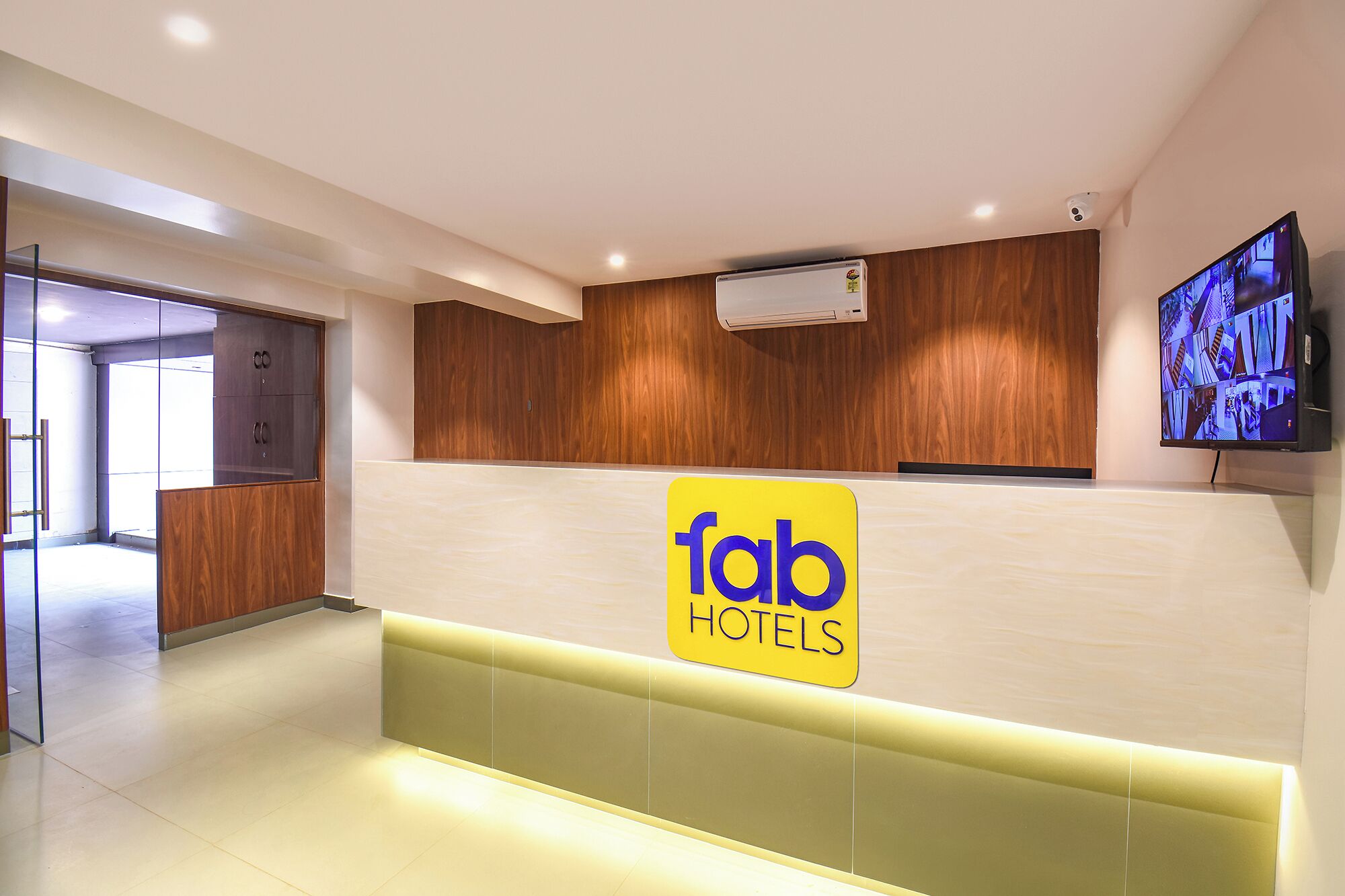 A reception area with a 'fab hotels' sign, wooden paneling, a flat-screen TV, and a glass door leading to another room.