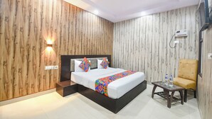 A modern bedroom with a wooden wall, a bed with a colorful geometric bedspread, a small table with water bottles, and a chair. - FabHotel Crown Residency - Near Lakhani Armaan Metro Station (Faridabad)