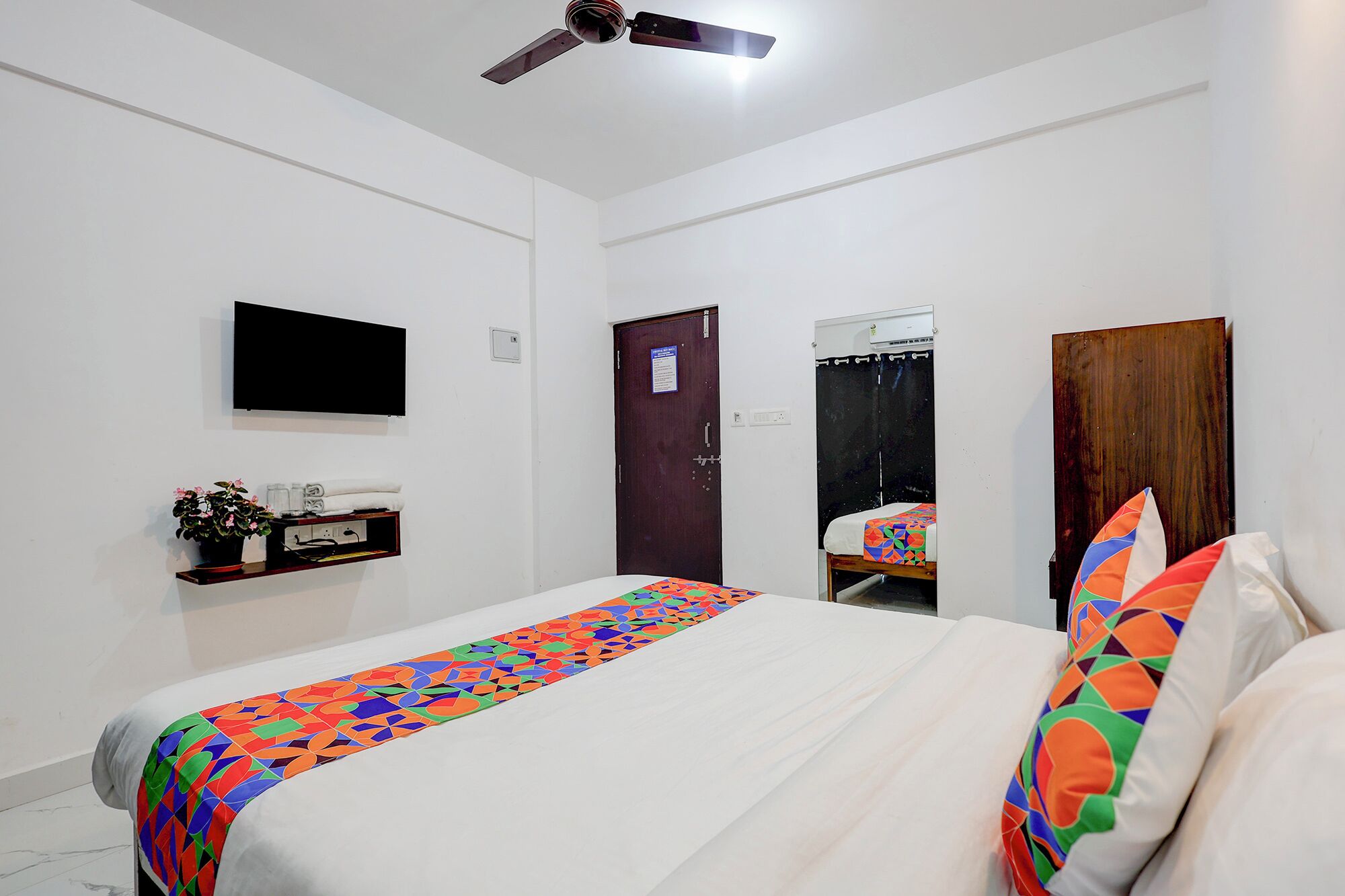 A bedroom with a bed, a TV mounted on the wall, a ceiling fan, a door, and a mirror.