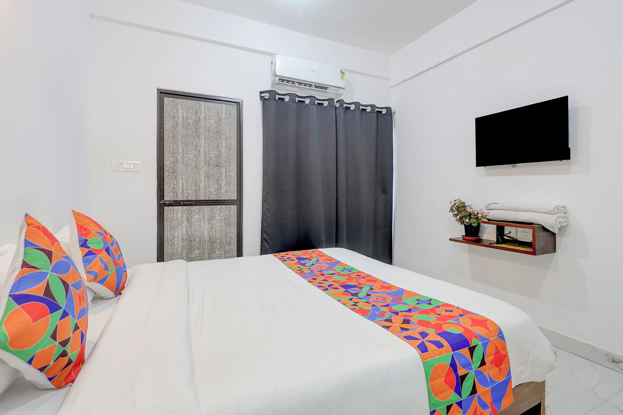 Een hotelkamer met een bed, een aan de muur bevestigde televisie, een klein plankje met een plant en een raam met gordijnen.