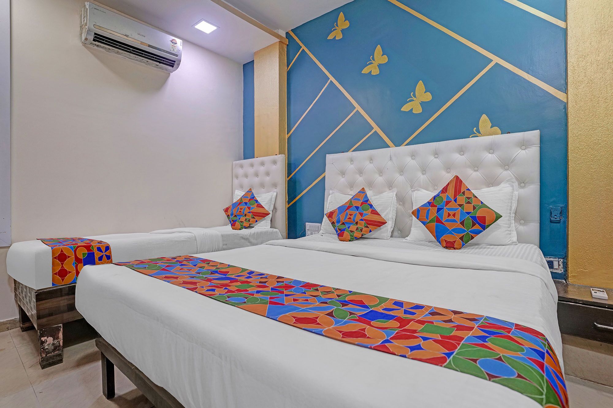 Ein Hotelzimmer mit zwei Betten, jeweils mit farbenfroher geometrischer Bettdecke und passenden Kissen. Das Zimmer verfügt über eine blaue Akzentwand mit goldenen geometrischen Mustern und einem weißen Wandgerät zur Klimatisierung.